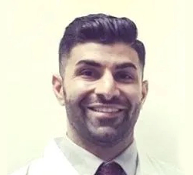 Professional headshot of smiling male dentist wearing white shirt and dark tie against neutral background.
