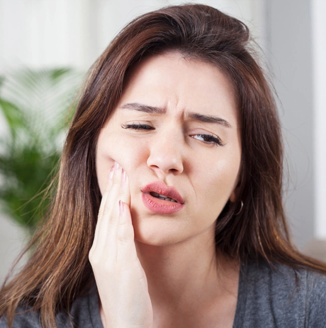 Woman holding cheek in pain, eyes closed, depicting dental discomfort or toothache indoors