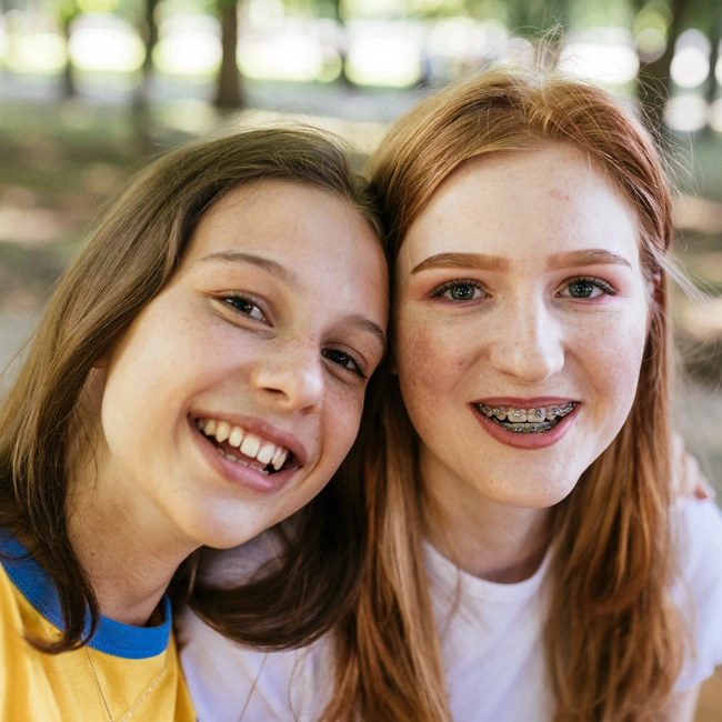 Two smiling teenage girls with braces posing together outdoors in a park setting.