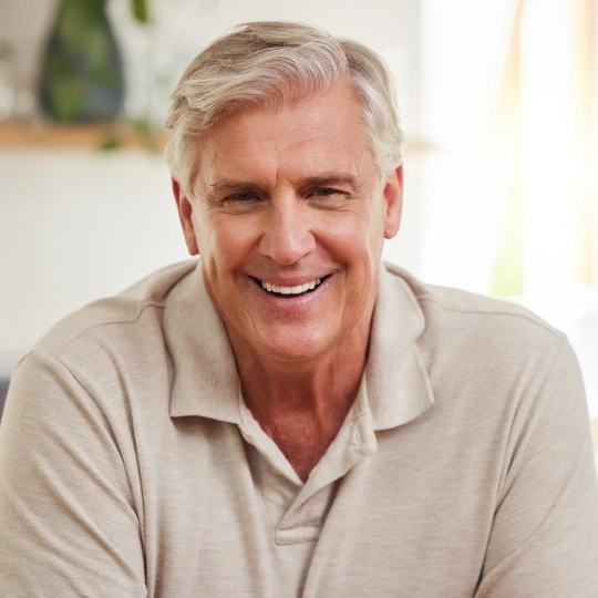 Smiling mature man in beige polo shirt, indoor home setting with blurred background.