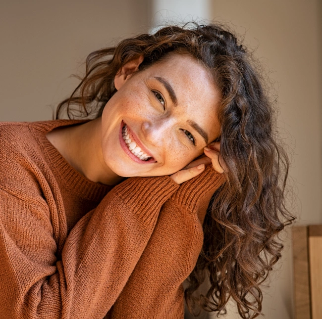 Smiling woman in rust-colored sweater leaning against shoulder in warm, neutral indoor setting.