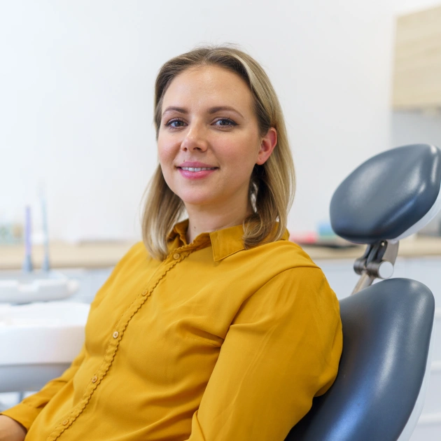 Female dental professional in yellow shirt seated in dental office chair, smiling at camera.