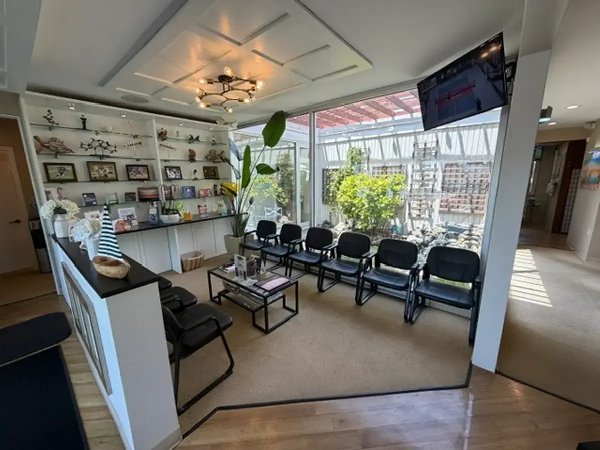 Wide-angle view of a bright dental office reception and waiting area, featuring a white front desk, black seating rows, a central coffee table, and large glass windows overlooking greenery.